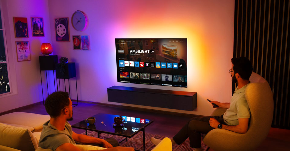 Zwei M&auml;nner schauen in einem Wohnzimmer auf einem an der Wand montierten Ambilight-Fernseher